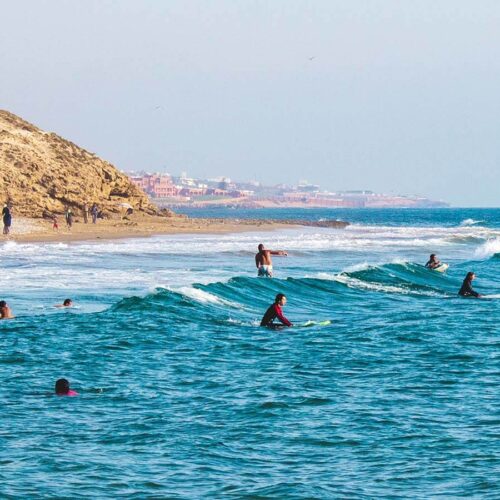 This captivating image depicts a group of people enjoying a Morocco tour, indulging in the thrill of surfing along the Atlantic. The scene is adorned with blue and white hues, reflecting the vibrant colors of their journey. Experience the luxury Morocco tour with Journeys Tours, where the exhilarating Morocco trip unfolds against a backdrop of teal waters and a bright blue sky. Immerse yourself in this dynamic scene, where surfers in wet suits ride the waves, while a car atop a hill offers a glimpse of adventure. Birds gracefully soar across the sky, adding a sense of movement to this mesmerizing Atlantic tour.