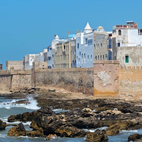 This image is of a stone wall with buildings on either side, overlooking the ocean in Essaouira (Morocco). The sky is grey and there are rocks along the shoreline. In the background, there is a large building that appears to be a castle or palace. There are also several smaller white cone-shaped buildings visible in the foreground.