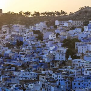 This image is a scenic view of Tangier in Morocco with many blue buildings on top of a hill. The sky is clear and the sun is shining, illuminating the white buildings in the foreground. There are several trees scattered around the hill, adding to its beauty. In the background, there are mountains visible in the distance.