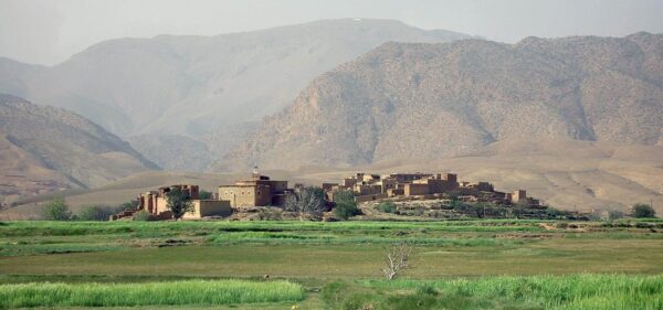 The Ait Bougumez valley in Morocco. This image is of a village in the desert. The scene is dominated by a large green field with buildings and mountains in the background. There are several trees scattered throughout the field, some with leaves and some without. In the foreground, there is lush grass growing close to the ground. A mosque can be seen on a dock in one corner of the image, while other small houses dot the landscape. The sky above is filled with fog or clouds, adding to its mysterious atmosphere.