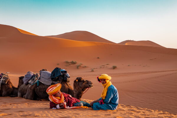 This image shows two Moroccan saharaians in the desert sitting on a sand dune in the desert, with three camels nearby. The sky is blue and there are dunes in the background. One of the people is wearing a yellow turban and a blue robe, while the other person has a colorful scarf wrapped around their head. There are also some clothes lying on top of a rock nearby.