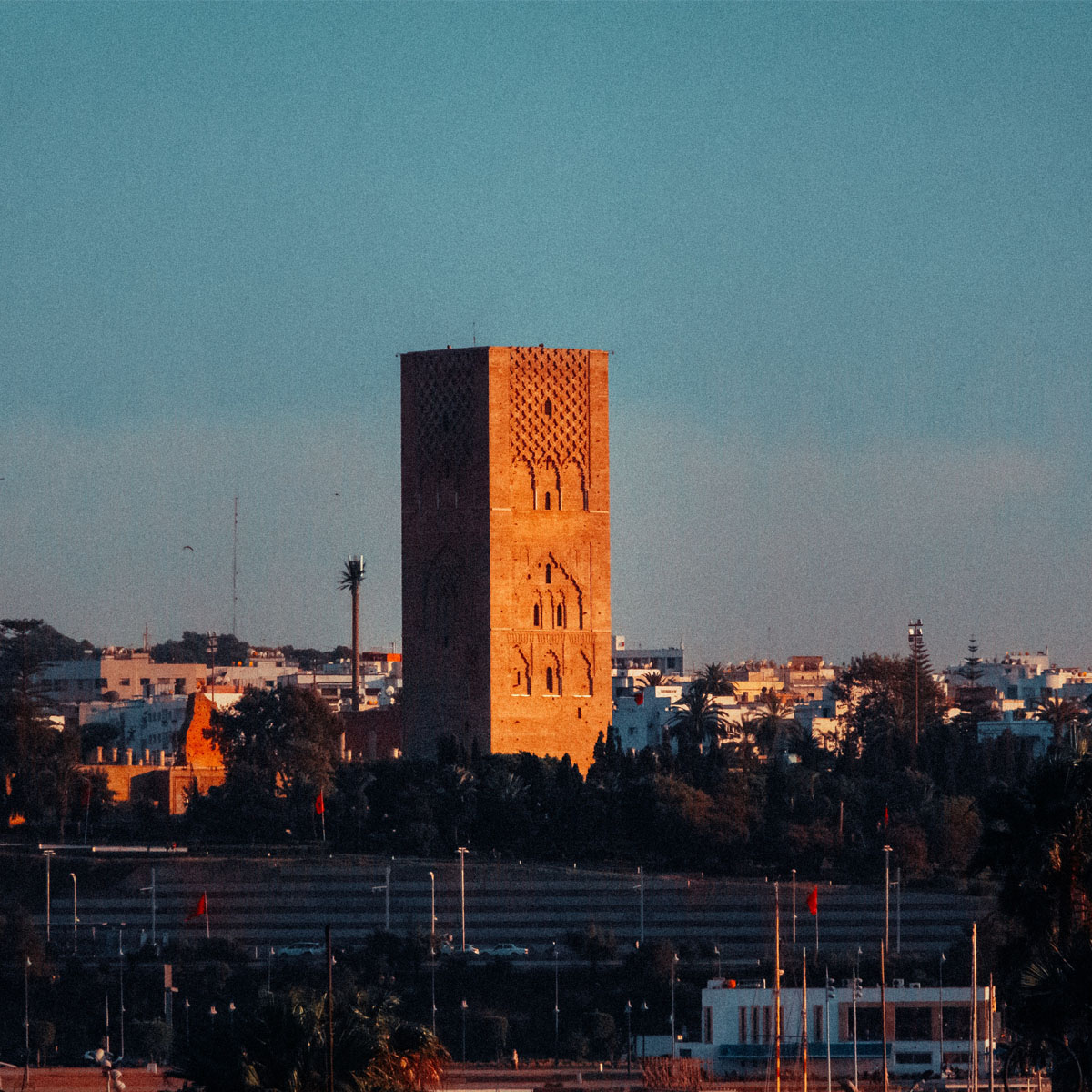 Marvel at the Magnificent Hassan II Tower in Rabat: A Symbol of Morocco's Rich Heritage. This image is of a tall brick tower in the middle of Rabat. The tower stands out against the blue sky and there are trees and buildings in the background.
